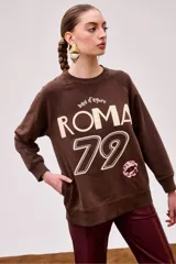 Buzo marrón con estampa frontal con la palabra "Roma" y el número 79 en color crudo. Tiene un parche con forma de boca en tonos burdeos.