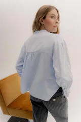 Camisa de algodón blanca con cuello a la base, cierre frontal con botones y mangas largas con puños delicados. Presenta volados verticales y detalles de cintas de terciopelo en contraste a lo largo del frente.