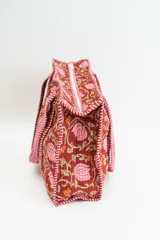 Bolso tipo tote acolchado con estampado floral en tonos rojizos, rosados y verdes. Presenta asas de mano con diseño de rayas verticales en color rojo y blanco, y ribetes a juego en los bordes.