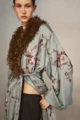 Kimono de largo midi en color verde grisáceo, con cuello de solapas amplias en pelo sintético marrón. Presenta mangas largas acampanadas, cierre frontal con cinturón ajustable del mismo tejido y un delicado estampado floral bordado en toda la prenda.