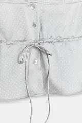 Top de tejido satinado color celeste con estampado de lunares blancos, escote en V con detalle de encaje beige, tirantes finos, cierre frontal con botones y lazo ajustable en la cintura.