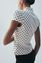 Top de popelín blanco con estampado de lunares negros, de manga corta, cuello redondo y cierre frontal con botones. Cuenta con bolsillos de parche en la parte delantera.