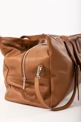 Bolso de viaje tipo weekender color marrón, confeccionado en cuero vacuno. Cuenta con doble asa de mano y correa bandolera ajustable y desmontable.