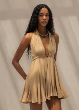 Vestido corto color beige con escote halter profundo, espalda descubierta y falda con vuelo. Presenta un diseño fluido y ligero con detalle de frunces en la cintura.