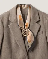 Pañuelo de seda con estampado de hojas y flores en tonos naranjas y grises sobre fondo beige, con bordes oscuros e incluye un dije dorado en forma de espiral.