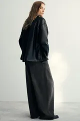 Camisa de efecto piel en color negro, con cuello solapa, manga larga y bolsillos de plastrón en el delantero. Presenta cierre frontal con botones ocultos por tapeta.