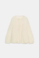 Blusa de viscosa color crema con cuello redondo, mangas largas abullonadas con puños elásticos y cierre frontal con lazadas. Presenta un diseño con bajo asimétrico y volantes.