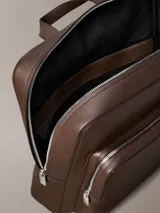 Mochila de hombre con silueta slim y cuadrada, confeccionada en material sintético con textura microgranulada (micro pebble). Color marrón oscuro, con asa de mano superior, correas acolchadas para el hombro, cierre de cremallera doble y bolsillo exterior frontal con cierre.