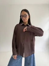 Camisa oversize de color marrón con cuello camisero y puños con detalle de bordado festoneado en color blanco. Presenta cierre frontal de botones y mangas largas.