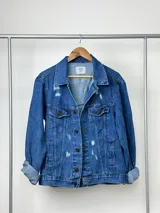 Campera de jean azul con cuello clásico, cierre con botones metálicos, bolsillos con solapa en el pecho y roturas en la parte delantera. Mangas largas con puños abotonados.