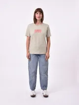 Remera de manga corta color beige claro con cuello redondo. Presenta un estampado gráfico central con un plato de pasta rodeado por un borde azul y blanco, sobre un fondo de franjas verticales rojas y blancas. El estampado incluye texto en cursiva en tonos turquesa y rojo.