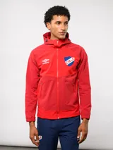 Campera deportiva Umbro del Club Nacional de Football, color rojo con capucha, cierre frontal y bolsillos laterales. Presenta el logo de la marca y el escudo del club en el pecho.