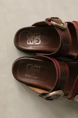 Sandalias bajas de cuero marrón con doble tira, hebilla ajustable y apliques de tachas metálicas.