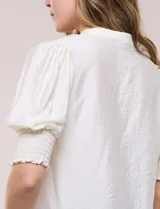 Blusa blanca de crepé con cuello mao y escote en V, cierre frontal con botones, mangas cortas abullonadas con puños elastizados en punto smock y calce semi holgado.