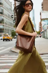 Bolso tipo saca de diseño asimétrico confeccionado en piel de color marrón. Cuenta con un asa de hombro ajustable y cierre mediante imán. Incluye una cartera con bolsillo extraíble en su interior.