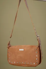 Cartera tipo baguette de gamuza color marrón, con correa ajustable y detalle de tachas metálicas plateadas en todo el contorno. Incluye placa metálica con logo en el frente.