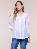 Blusa blanca de silueta suelta y textura liviana, con escote en V, cuello mao y cierre frontal de botones. Cuenta con alforzas verticales en el pecho y mangas tres cuartos con puños elastizados y detalle de galón calado.