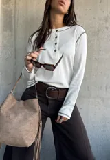 Remera de manga larga en color blanco, confeccionada en tejido acanalado. Presenta un diseño tipo henley con cuello redondo y una pequeña botonadura frontal, destacada por costuras a contratono en color oscuro que recorren los hombros y los puños.