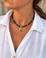 Collar de cristalitos facetados en color verde oscuro con cuentas metálicas doradas intercaladas, y un dije ovalado de acero con baño en oro y detalle de piedra brillante incrustada.