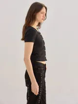 Remera negra de canalé ajustada al cuerpo, de manga corta y cuello redondo, con cierre frontal de botones metálicos.