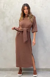 Vestido largo color verde militar, con cuello redondo, mangas largas abullonadas con puños elásticos y detalle de lazo anudado en la cintura. Presenta una abertura lateral en la falda.