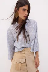 Camisa de algodón color azul claro con rayas verticales blancas y bordado calado de flores blancas. Tiene cuello clásico, cierre frontal con botones y mangas 3/4 acampanadas.