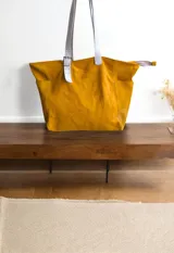 Bolso tipo tote de cuero color mostaza con asas de cuero sintético color gris claro. Tiene dos bolsillos externos y cierre superior con cremallera.