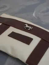 Sobre tipo cartuchera de lona naranja con detalles y apliques en cuero marrón. Presenta un aplique metálico en forma de caballo y un parche con el logo de la marca grabado en el frente.