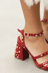 Sandalias de tacón ancho de 7 cm, color bordó, con diseño de tiras cruzadas en el empeine y pulsera al tobillo con cierre de hebilla. El modelo está decorado con tachas metálicas plateadas y doradas distribuidas por todas las tiras y el tacón.