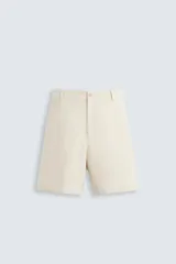 Bermuda de corte relajado color beige claro, confeccionada en tejido con textura. Presenta cierre frontal con cremallera y botón, bolsillos delanteros y bolsillos traseros de doble vivo con botón.