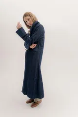 Tapado largo de jean azul oscuro, con cuello solapa y puños con detalle de cinta de terciopelo negro. Presenta costuras visibles que definen paneles y un logo bordado en la espalda.