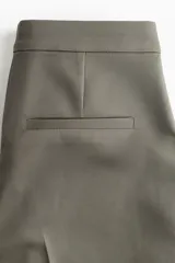 Pantalón cigarette tobillero color verde militar, con cintura estándar, cierre y pretina prolongada con corchete oculto. Presenta piernas ajustadas con abertura corta en los bajos, bolsillos al bies y un bolsillo decorativo en la parte trasera. Incluye pliegues en la parte delantera y trasera.