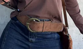 Faja ancha de gamuza color camel con tachas metálicas doradas en los bordes y hebilla circular grande de metal envejecido.