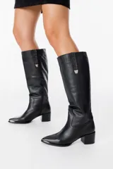 Bota bucanera de cuero negro con taco cuadrado y punta fina. Presenta un detalle decorativo vertical con tachas metálicas en la parte superior de la caña y puntera metálica.