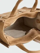 Cartera tipo tote bag de lona color beige, con diseño estructurado y asas superiores. Presenta el logo de la marca estampado en la parte frontal y detalles de cintas laterales con inscripciones.