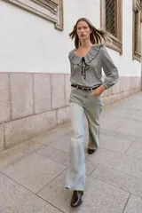 Blusa de manga larga con estampado de cuadros vichy en blanco y negro. Presenta un escote en V adornado con un volante amplio y un lazo decorativo en el centro. Las mangas terminan en puños elásticos.