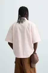 Camisa de algodón color rosa pálido, corte boxy fit, con cuello solapa, manga corta y abotonadura frontal. Presenta un texto bordado en contraste de color rojo en el delantero.