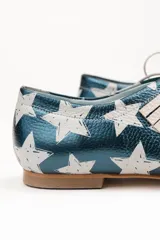 Zapato tipo blucher de cuero con acabado metalizado en color azul petróleo y estampado de estrellas blancas. Presenta un diseño con flecos decorativos en la parte frontal, cierre con cordones y suela plana de cuero.