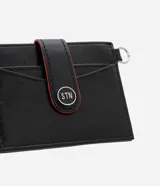 Billetera compacta de poliuretano color negro, con solapa frontal con detalles en rojo, botón a presión metálico con logo, compartimentos para tarjetas y argolla lateral.