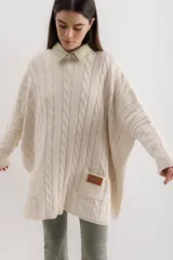 Sweater de punto color camel con diseño de trenzas verticales y acabado brillante. Presenta una silueta holgada tipo poncho, cuello redondo, mangas largas con puños ajustados y bolsillos frontales con detalle de etiqueta de cuero.