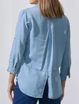 Camisa de tencel color celeste claro, con cuello con solapa, escote en V y abertura central con botones. Manga larga con presilla para ajustar el largo. El largo de espalda es mayor al delantero y tiene un tajo central.