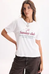 Camiseta blanca de manga corta con estampa frontal en color bordó y verde con la leyenda 'The Sunday Club' y texto descriptivo.