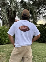 Remera blanca de algodón con estampado de un balón de fútbol americano y la frase "Together, we are America" en rojo. Incluye una firma en negro debajo del estampado.