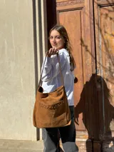 Cartera tipo tote bag de cuero color camel, con detalles de tachas metálicas en el bolsillo frontal y tiras largas para el hombro.