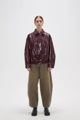 Chaqueta de eco-cuero en color bordó con acabado brillante, diseño holgado, cuello camisero, cierre frontal con botones a presión y detalle de elástico fruncido en el ruedo.