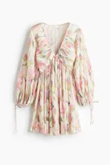 Vestido corto con textura plisada, estampado floral en tonos pastel (rosa, verde, amarillo, azul) sobre fondo crema. Presenta escote pronunciado en V con fruncido central y mangas globo largas ajustadas con lazos en los puños.