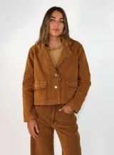 Chaqueta corta de pana color tostado, con cuello solapa, cierre frontal de botones y bolsillos delanteros con solapa. Presenta detalles de bordes deshilachados en el cuello, solapas y bolsillos.