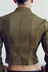 Chaqueta corta de antelina color verde militar, con cuello subido, manga larga y bajo asimétrico. Presenta detalles de alamares negros bordados en contraste en el frente y en los puños.