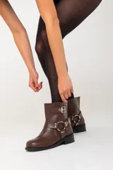 Botas cortas de ecocuero color chocolate, con base de 1,5 cm, taco de 2,5 cm y caña de 21 cm de alto. Presentan dos correas con hebillas y un arnés con argolla metálica.