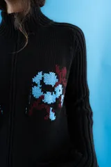 Cardigan de punto color marrón con cierre frontal, cuello alto y bolsillos aplicados. Presenta diseño tejido localizado con detalles en violeta y negro en el pecho.